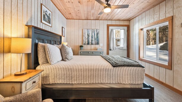 A bedroom with a bed, nightstand, lamp, ceiling fan, artwork, and a window with a view of trees.