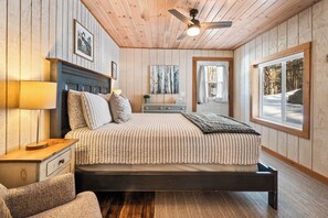 A bedroom with a bed, nightstand, lamp, ceiling fan, artwork, and a window with a view of trees.