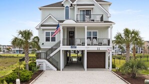 A two-story house with a balcony, a garage, and a flag. - Marshfront Home w/ Private Pool, Elevator, & Easy  Access by Williamson Realty (Ocean Isle Beach)