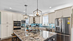 A modern kitchen with white cabinetry, granite countertops, stainless steel appliances, and pendant lighting. - Marshfront Home w/ Private Pool, Elevator, & Easy  Access by Williamson Realty (Ocean Isle Beach)