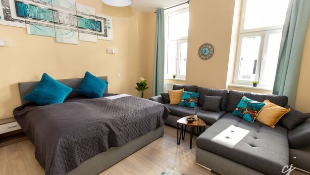 A modern bedroom with a bed, a sofa, and a small table. There are curtains on the windows, and a clock is visible on the wall. - Shimmering Blue | By Radiant Apartments (Wien)