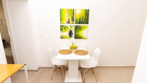 A dining area with a white table and two white chairs, a small wooden table to the left, and a wall with four framed paintings. - Shimmering Blue | By Radiant Apartments (Wien)