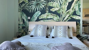 Un lit avec des coussins à motifs, une tête de lit en bois et un papier peint orné de motifs tropicaux.