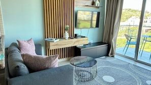 Un salon moderne avec un canapé, une table basse, un téléviseur fixé sur un panneau en bois et une vue sur l’extérieur.