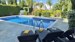A swimming pool with a tiled edge, surrounded by stone flooring, and two wicker chairs with towels.