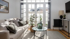 A modern living room with a sofa, a glass coffee table, and a view through large windows. - Beautiful Mews Cottage | 6 min walk to Rottingdean Beach | Near Brighton (Rottingdean)