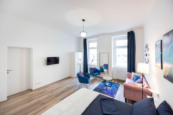 Quarto de hotel moderno com cama, escrivaninha, cadeira e sofá. Há uma televisão na parede e uma janela com cortinas.