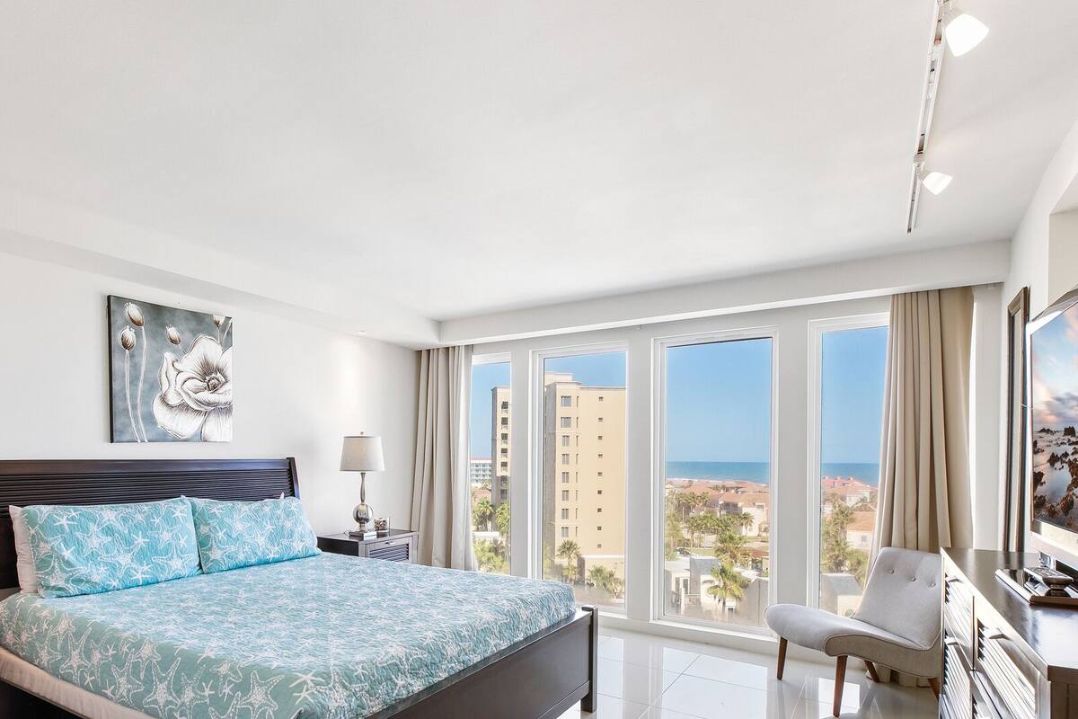 A modern hotel room with a large bed, two bedside tables, a desk with a chair, a large window with a city view, and a painting on the wall.