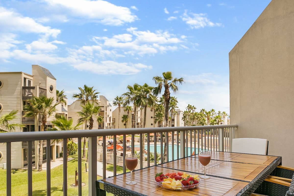 A balcony with a table set for two, overlooking a pool area with palm trees and a clear sky.