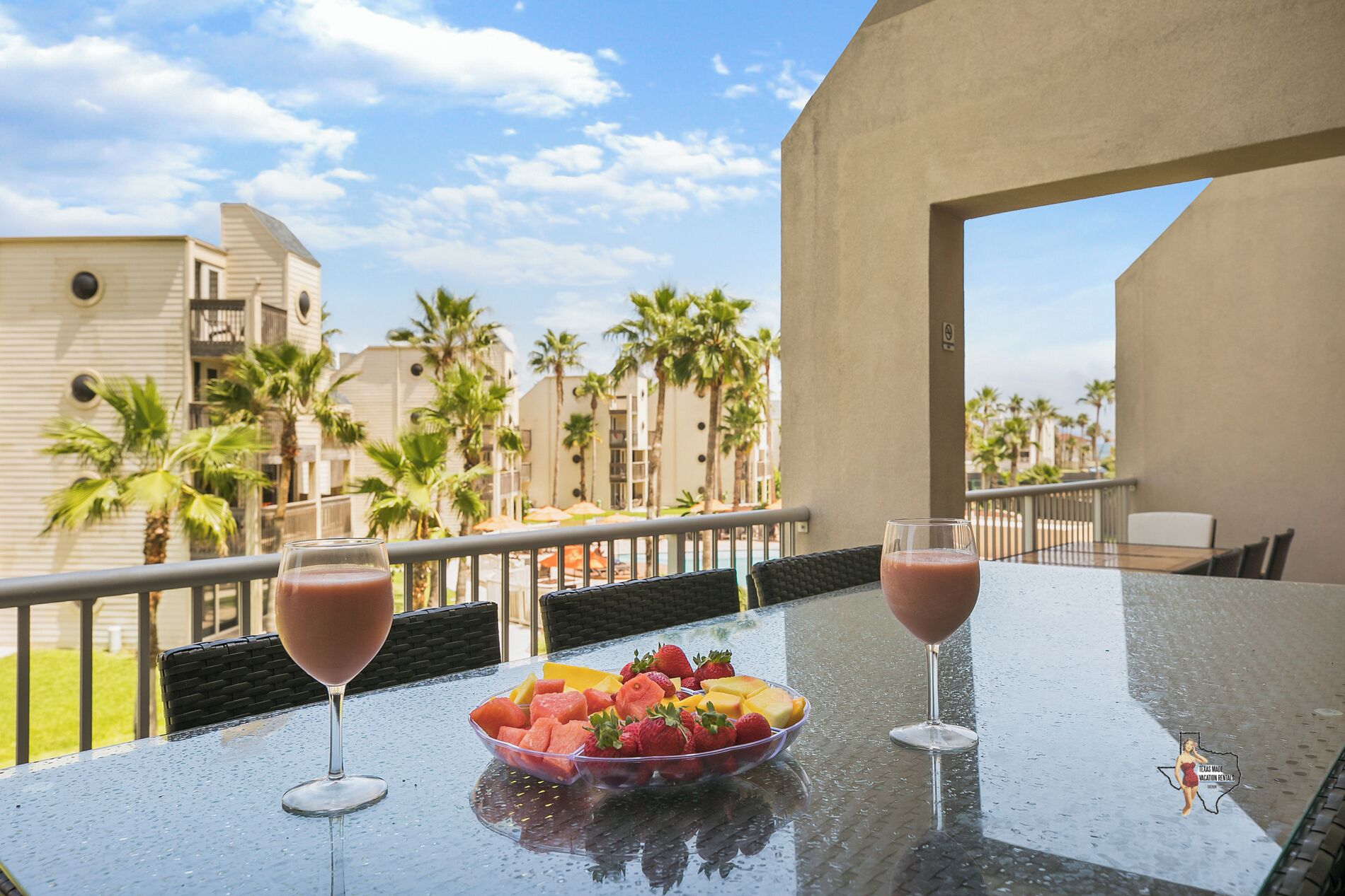 A balcony with a table set for two, featuring a fruit bowl and two glasses of a pink beverage.