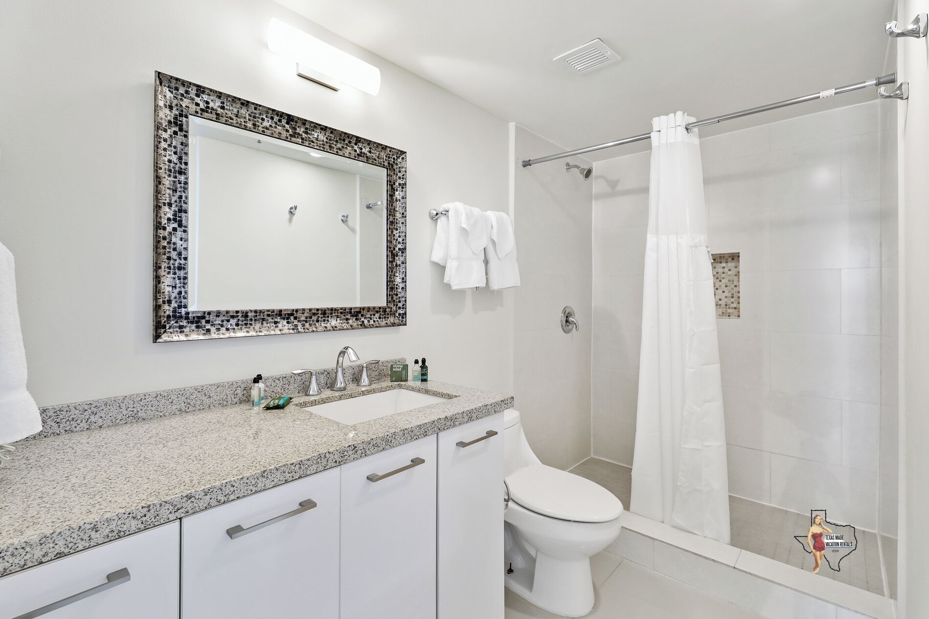 Ein Badezimmer mit einer Granittheke, einer weißen Toilette, einer Dusche mit Glastür und einem Spiegel mit Mosaikrahmen.