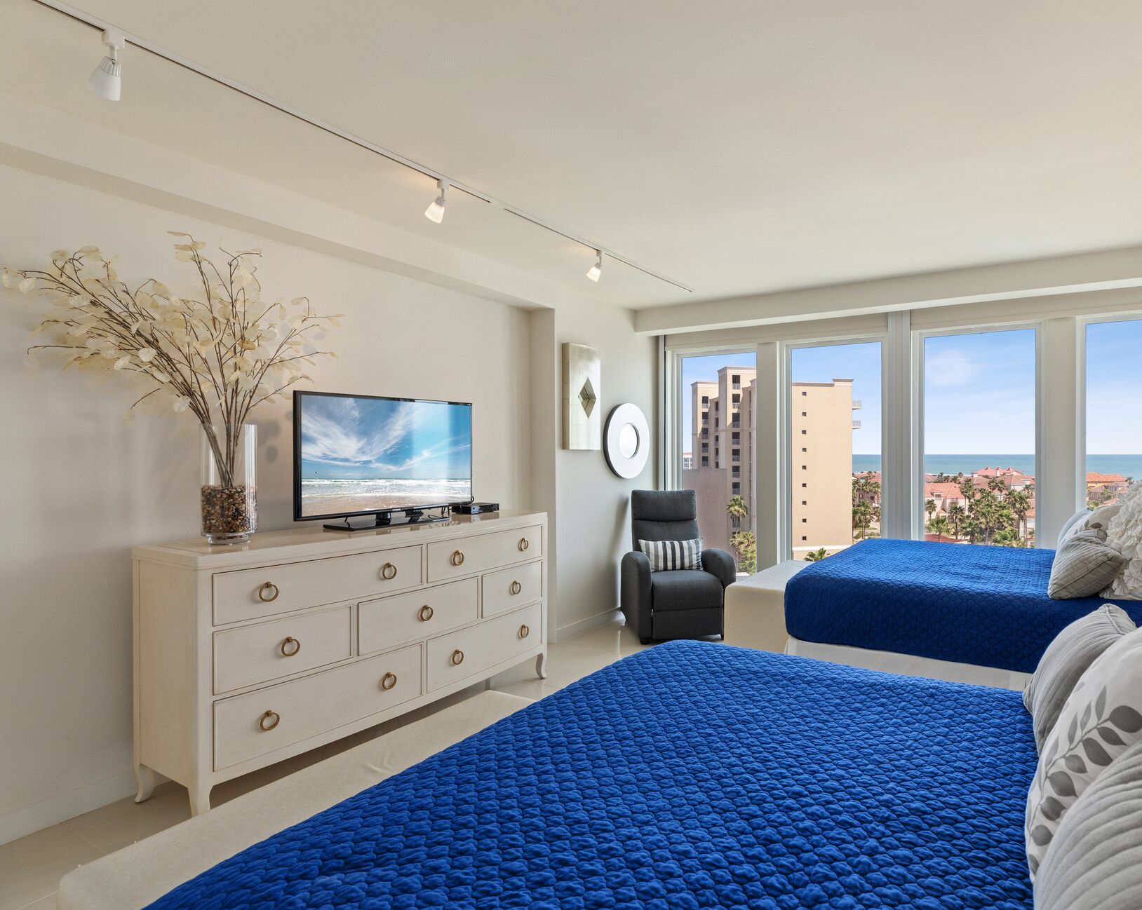Une chambre d’hôtel moderne dotée d’une grande fenêtre, d’un téléviseur à écran plat, d’une commode blanche, de deux lits avec une literie bleue et d’un vase contenant des fleurs séchées.