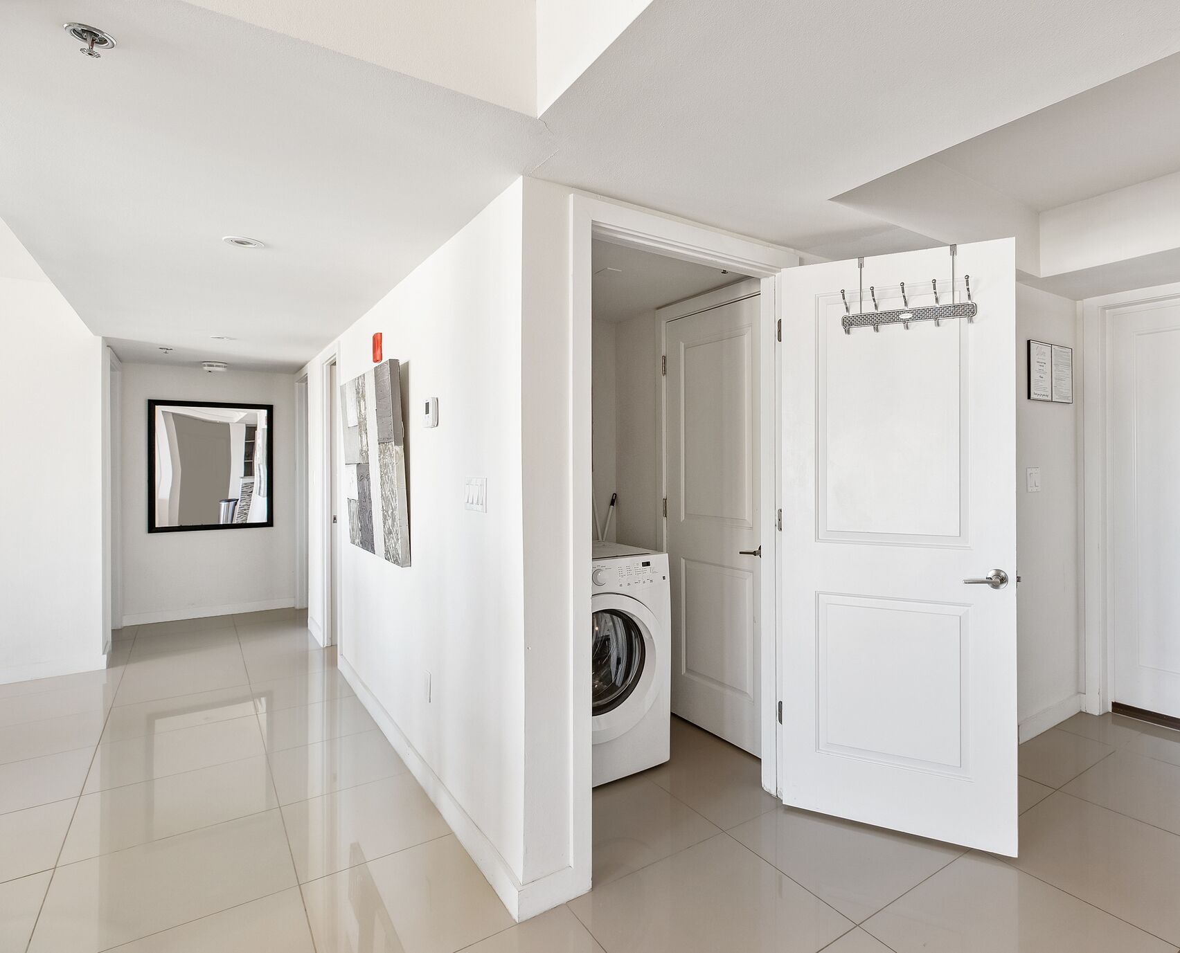 Un pasillo moderno y amplio con lavadora, una puerta con perchero y un espejo que refleja la habitación.