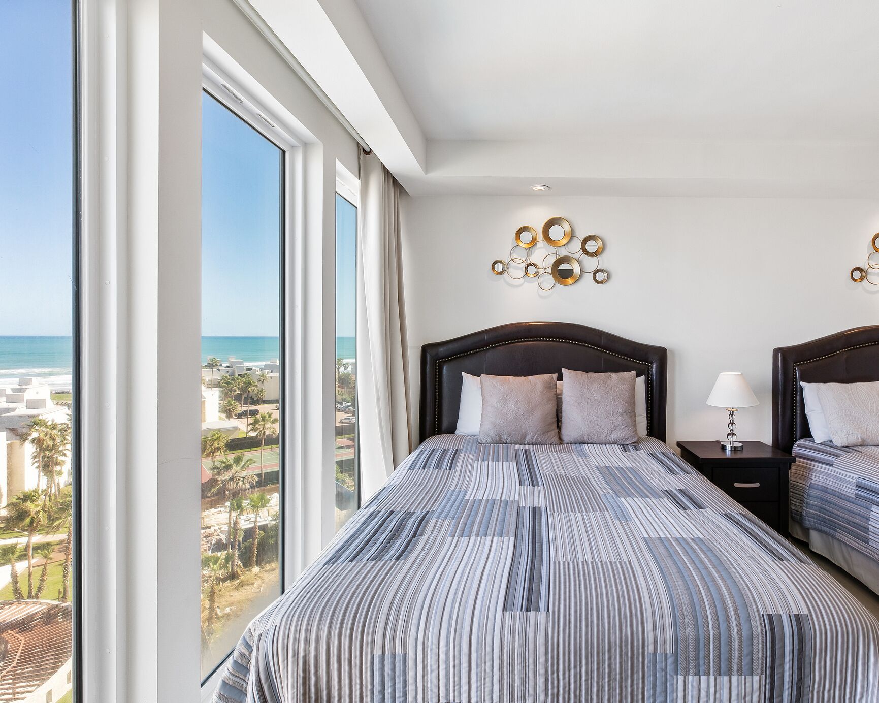 Una camera da letto con un'ampia finestra vista spiaggia, un letto con lenzuola a righe, un comodino con una lampada e un quadro appeso al muro.