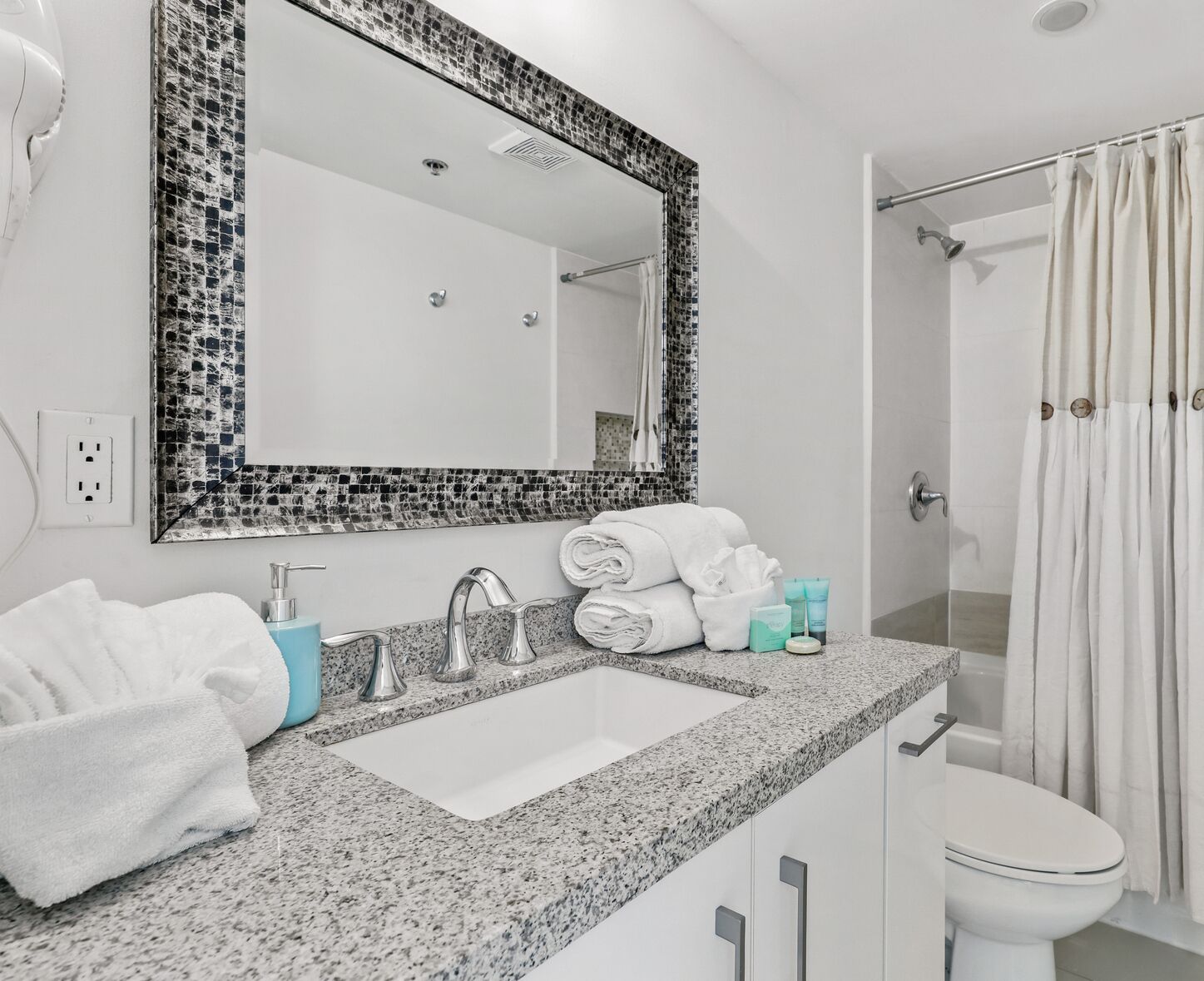 Un bagno con lavabo su piano in granito, un ampio specchio con cornice a mosaico, un water bianco e una doccia con tenda.