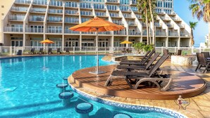 Un espace piscine d’hôtel avec des chaises longues, des parasols et un immeuble à plusieurs étages en arrière-plan.