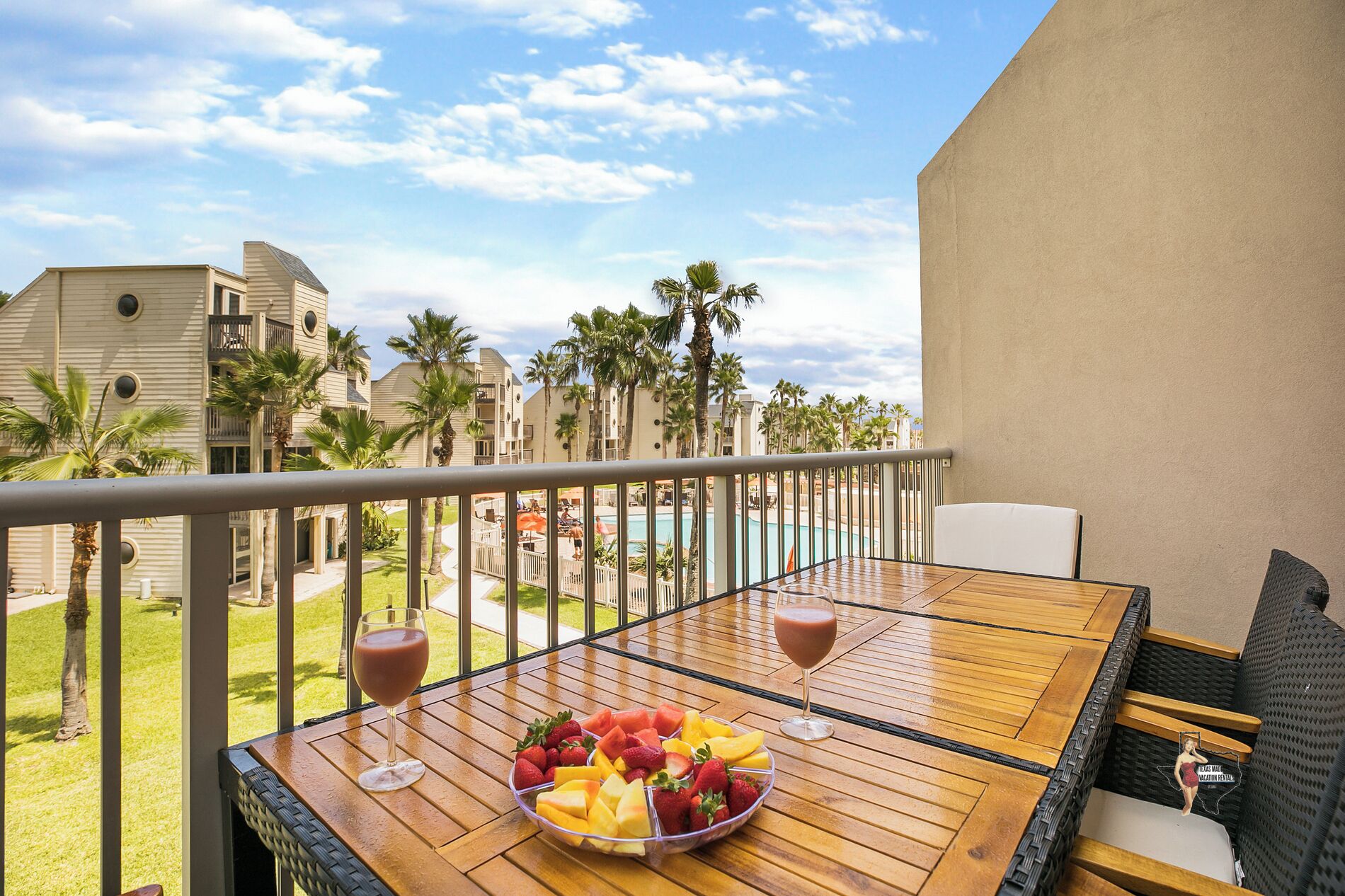 A balcony with a wooden table, two glasses of red beverage, and a fruit bowl.