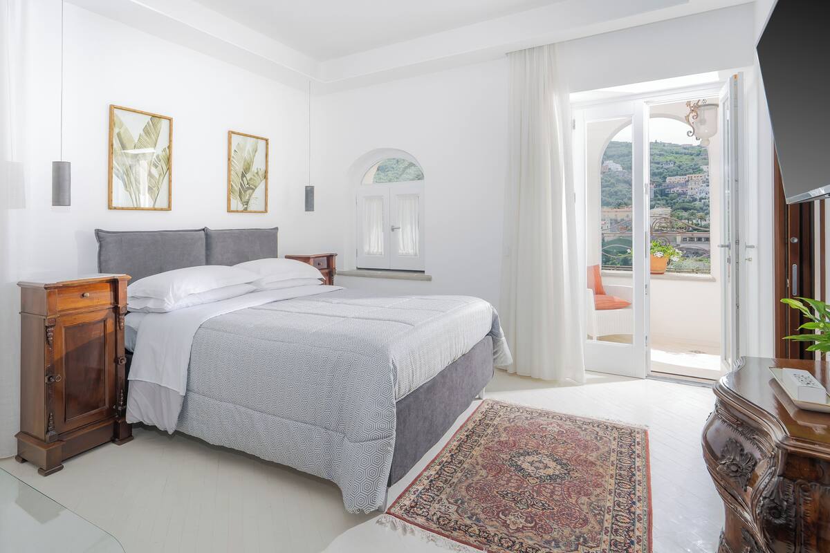 Une chambre à coucher comprenant un lit, une table de chevet, une commode, une fenêtre avec des rideaux et une vue sur un balcon agrémenté d’une plante.