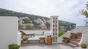 A rooftop terrace with a view of a hillside town, a wooden bench, potted plants, and a cactus. - Le Cavottole Resort (Vico Equense)
