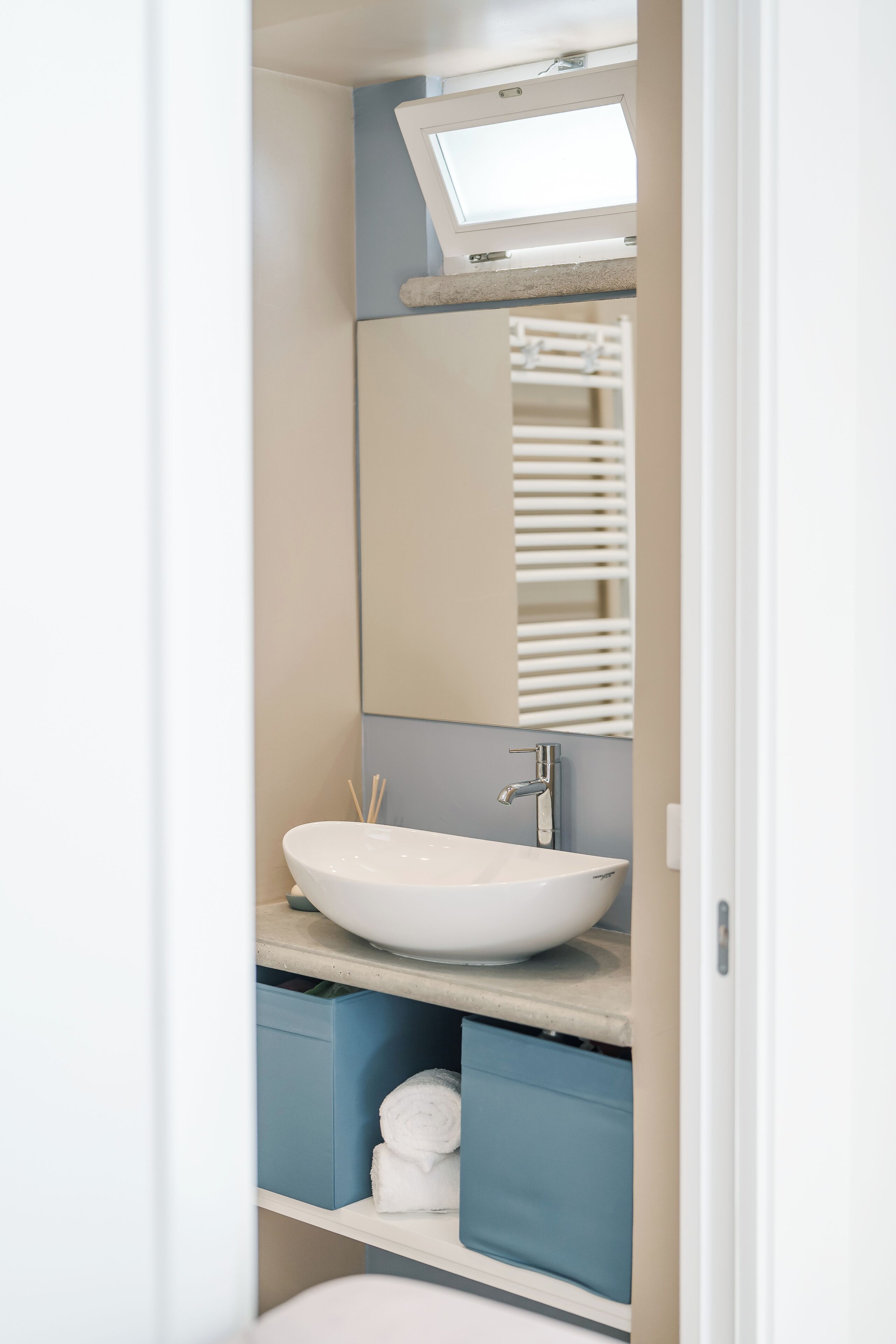 Une salle de bain avec un lavabo blanc, un miroir et une fenêtre au-dessus du lavabo. On trouve des tiroirs bleus sous le lavabo, qui servent de rangement.
