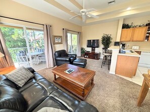 A living room with a leather sofa, a wooden coffee table, a TV, and a kitchen area with a countertop and cabinets.