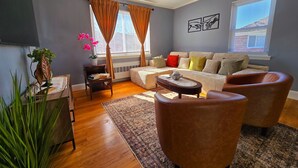 A living room with a sofa, two armchairs, a coffee table, a side table with a plant, and a rug. - Under the Sun Apartment (Boston)