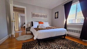 A bedroom with a bed, a leopard print rug, a window with curtains, and a hallway leading to another room. - Under the Sun Apartment (Boston)