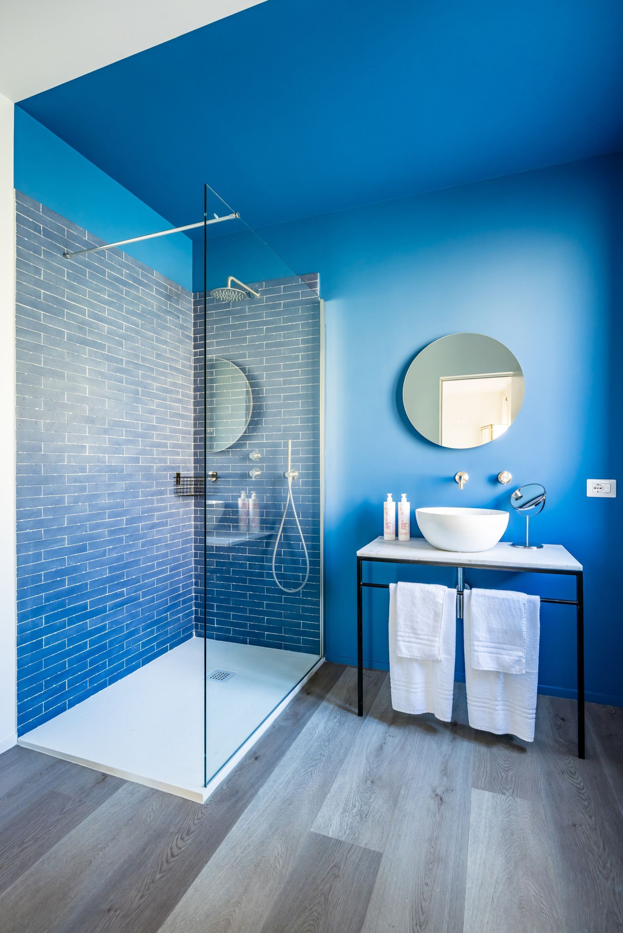 A bathroom with a glass-enclosed shower, a white sink, and a round mirror on a black countertop.