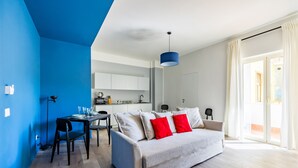 A modern living room with a sofa, dining table, and chairs. There is a kitchen area in the background with cabinets and a countertop. - Street Stritt 60 by Wonderful Italy - Apt 2 (Naples)