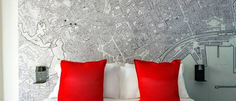 Une chambre d’hôtel avec une carte détaillée de la ville accrochée au mur, un lit avec des oreillers blancs et rouges, et une table de chevet avec une lampe.