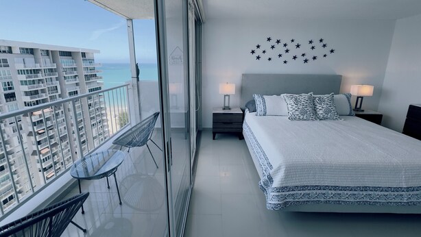 Une chambre dotée d’un grand lit, de deux tables de chevet avec des lampes, d’un balcon avec une chaise, et offrant une vue sur la mer et les bâtiments.