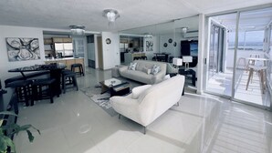 Un salon moderne avec un canapé, une table basse et un coin repas. On aperçoit une cuisine au fond, et un balcon avec vue.