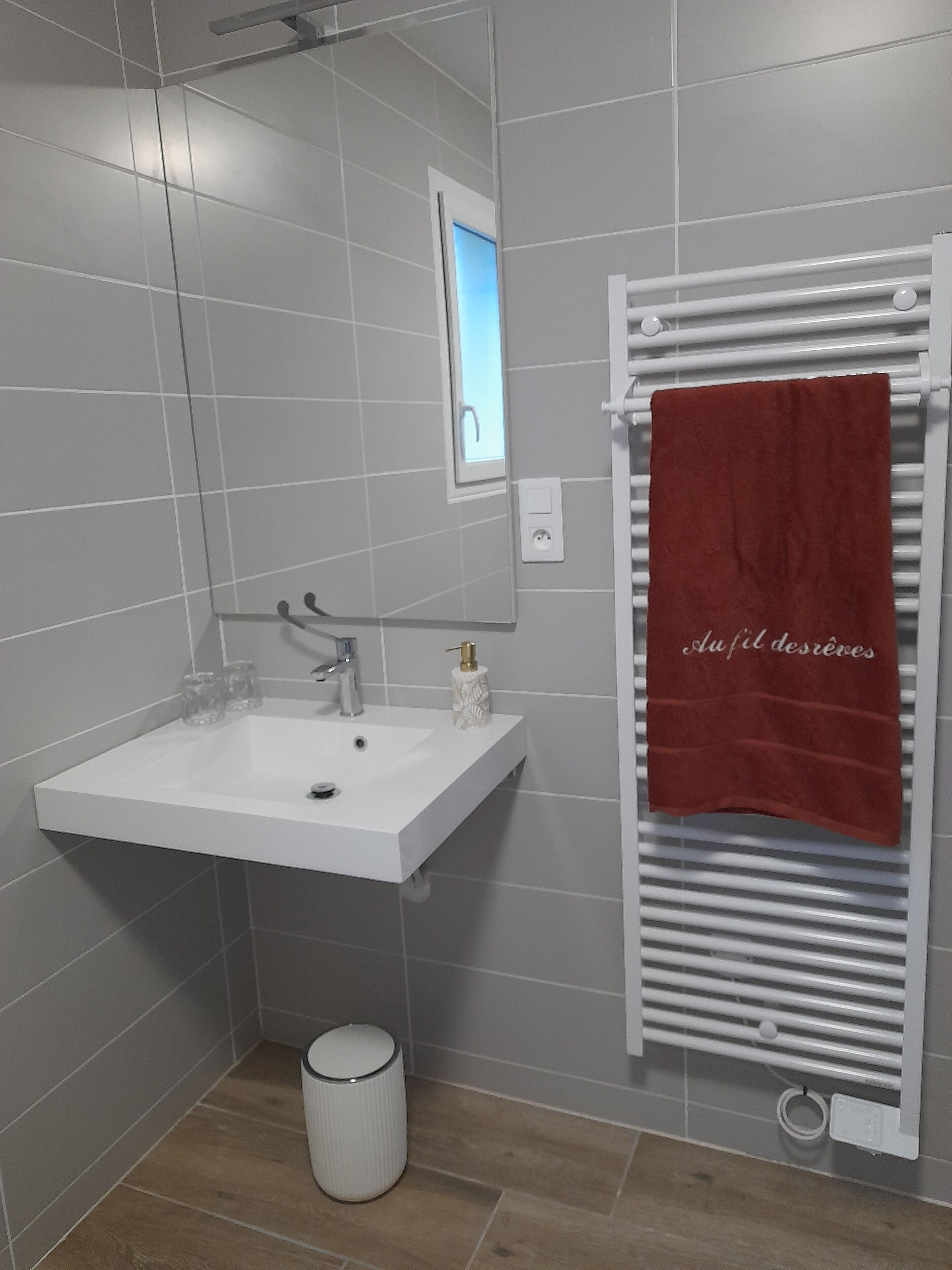 Une salle de bain avec un lavabo blanc, un miroir, un porte-serviettes avec une serviette rouge et une poubelle.