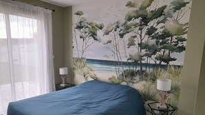 Een slaapkamer met een bed, twee nachtkastjes met lampen, een groot wanddecoratie van een strandgezicht, en dunne gordijnen voor het raam.