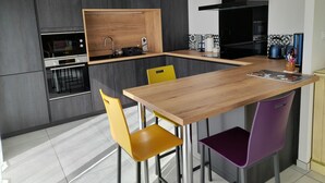 Een moderne keuken met een houten aanrecht, een inbouwoven en een spoelbak. Er staan twee stoelen, een geel en een paars, en een zwarte afzuigkap boven het fornuis.