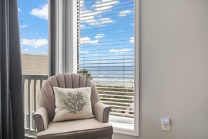 A chair with a cushion, a window with blinds, and a view of the ocean.