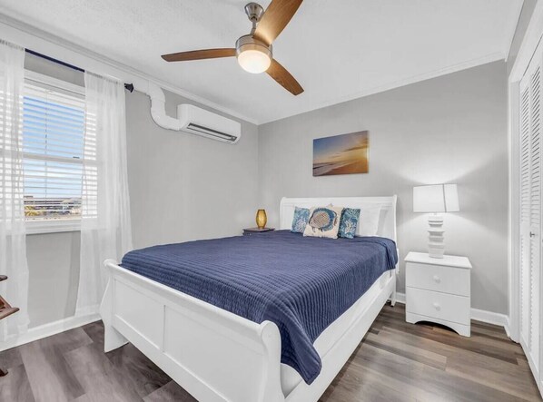 Un dormitorio con una cama, una mesita de noche, un ventilador de techo y una ventana con cortinas.