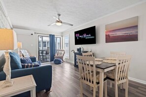 Una sala moderna con un sofá, una mesa de comedor, sillas, un ventilador de techo y un televisor que muestra 'Sunny Beach Vacation Rentals'.