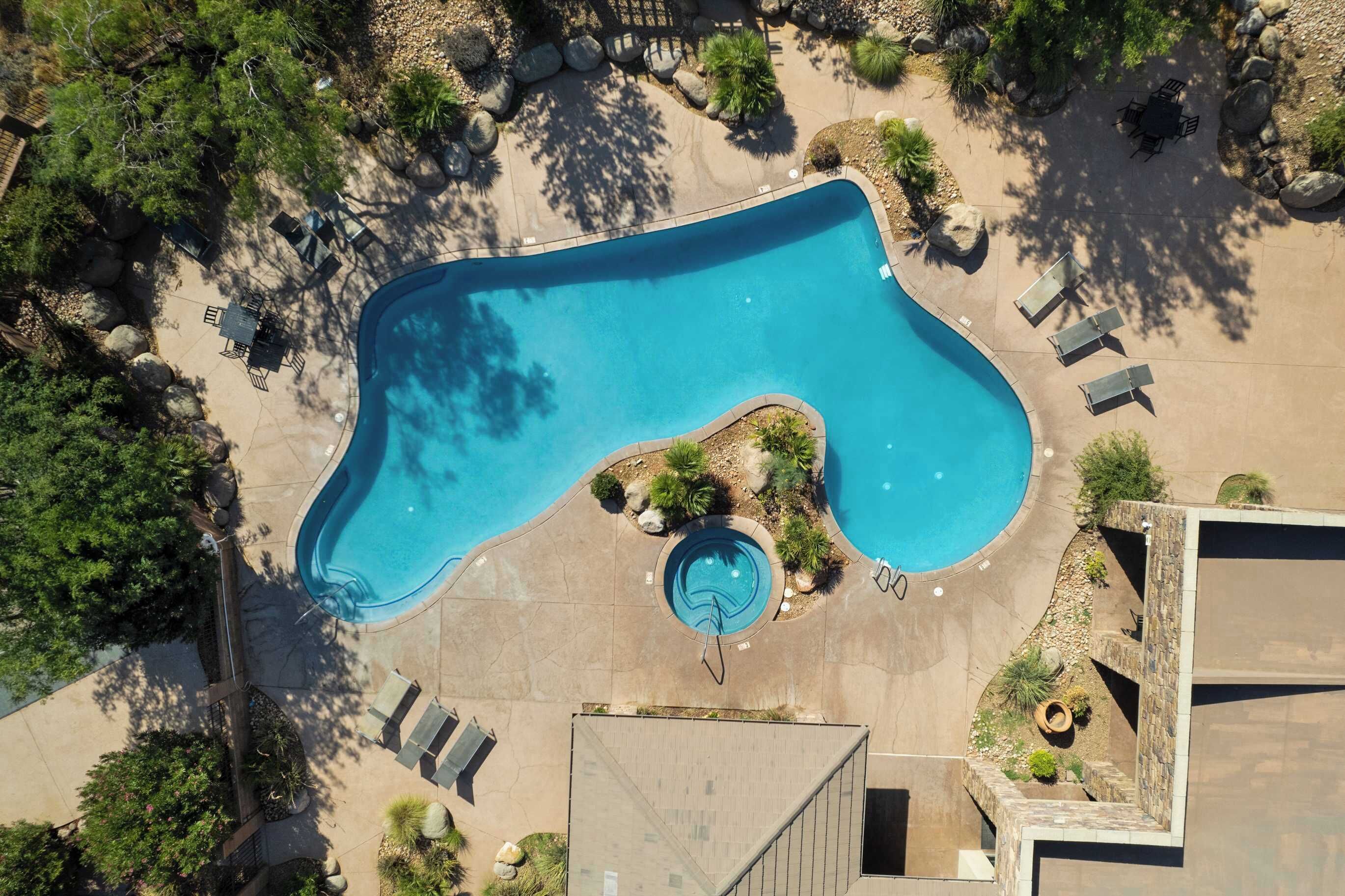An aerial view of a swimming pool surrounded by lounge chairs and a small hot tub, with trees and rocks in the background.