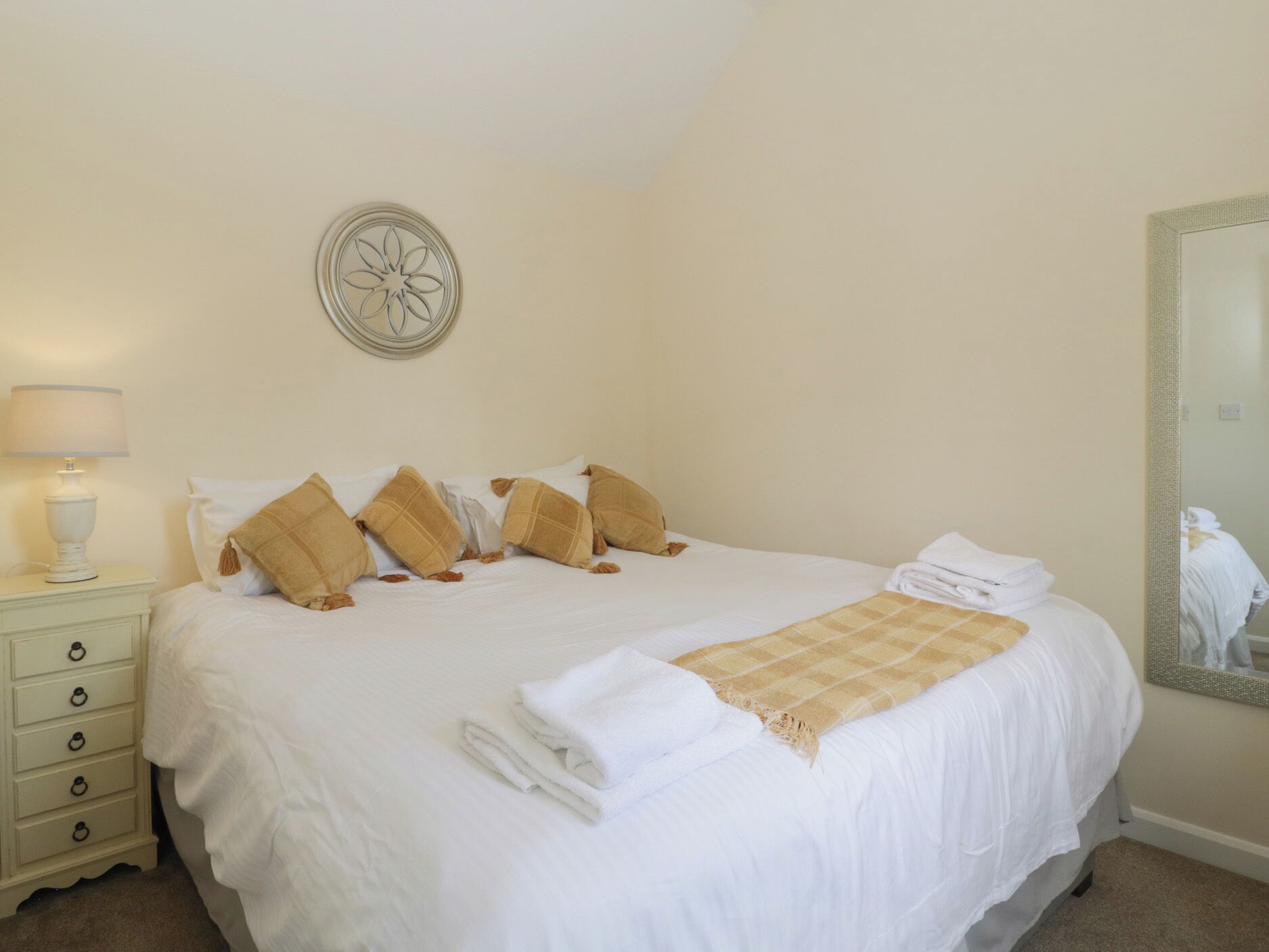 A neatly made bed with white linens and a beige throw blanket, a bedside lamp, and a wall-mounted mirror.