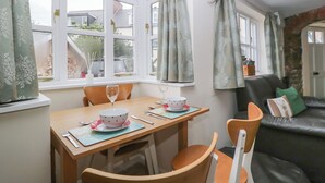Un coin repas avec une table en bois, des chaises et un ensemble de bols. On y trouve des fenêtres avec des rideaux, un mur en pierre et un canapé en cuir noir agrémenté de coussins.