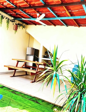 Ein überdachter Patio-Bereich mit Deckenventilator, Kühlschrank und Holztisch.