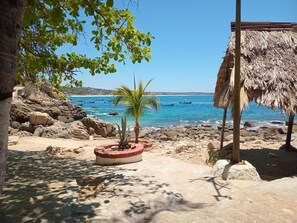 Ein Strand mit einer strohgedeckten Struktur, einer Palme und einer felsigen Küstenlinie.