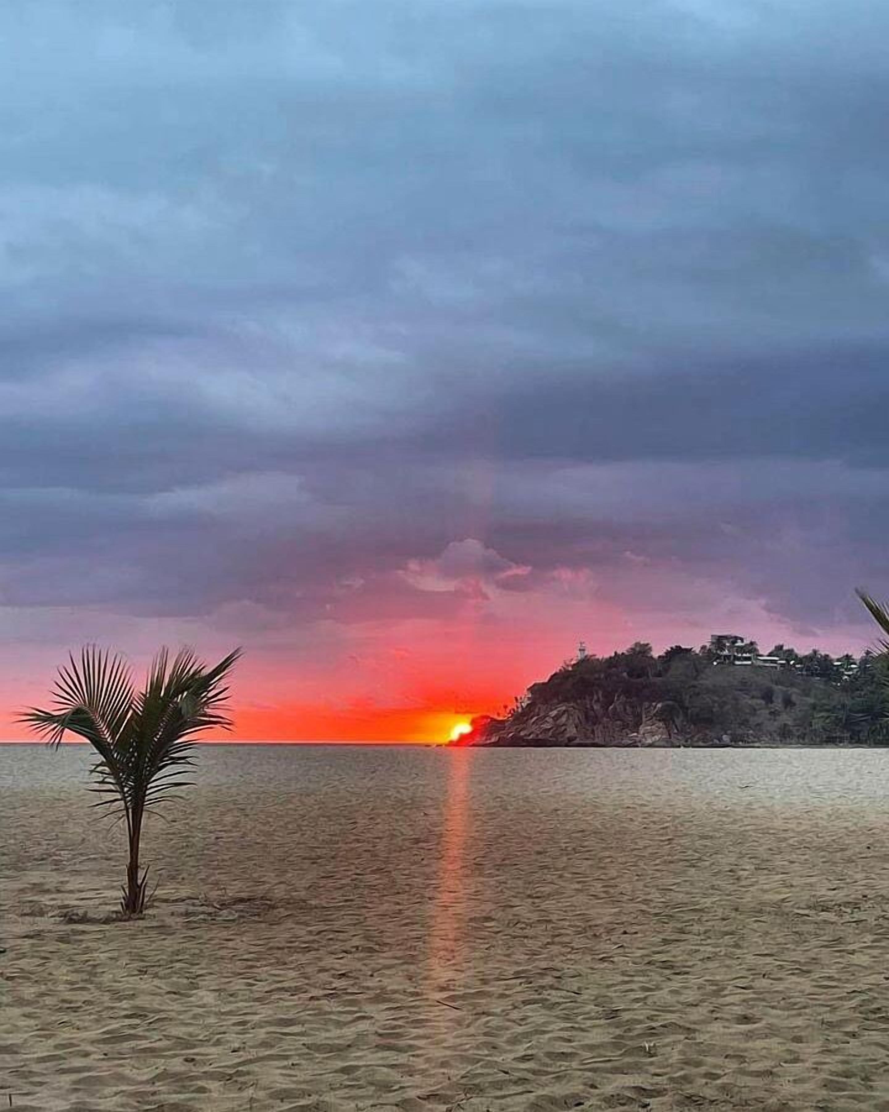 Una playa con atardecer, palmeras y un promontorio rocoso a lo lejos.