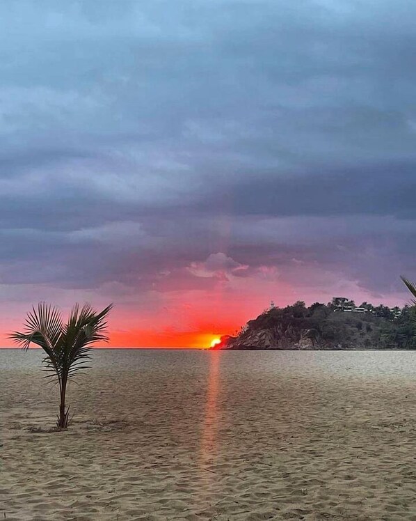Una playa con atardecer, palmeras y un promontorio rocoso a lo lejos.