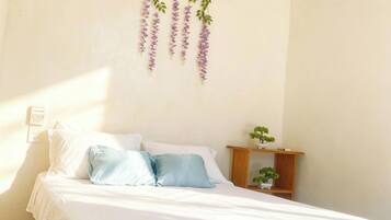 Un dormitorio con cama, ropa de cama blanca, un equipo de aire acondicionado, una planta colgante y una mesita de noche de madera con una planta.