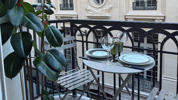 Un balcon avec une table dressée pour deux personnes, des plantes en pot et une vue sur un bâtiment classique aux fenêtres richement décorées.