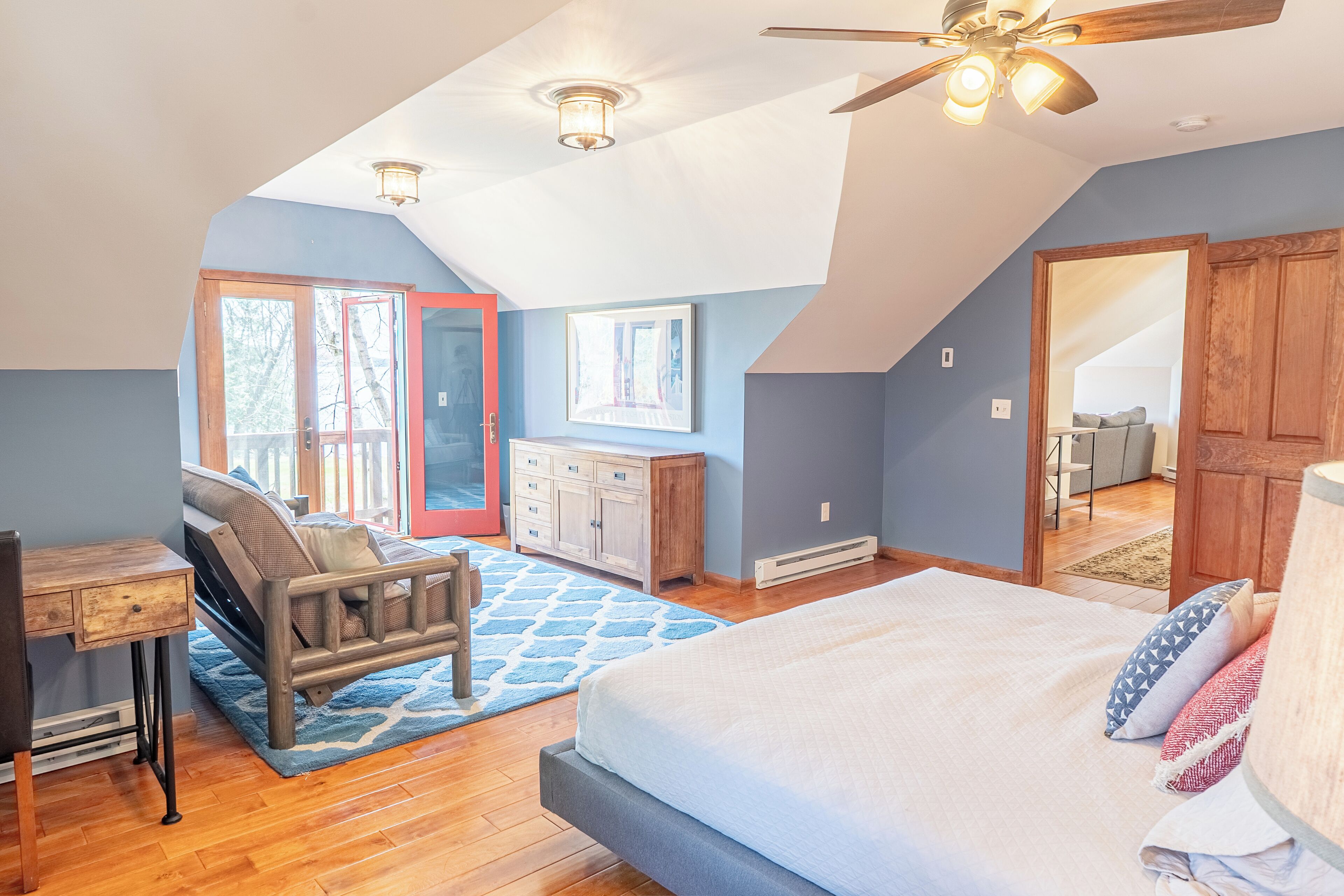 A bedroom with a large bed, a desk, and a chair. There is a ceiling fan, a window, and a door leading to another room.