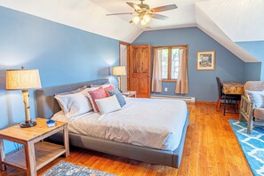Ein Schlafzimmer mit Bett, Nachttischen aus Holz, einem Deckenventilator, einem Fenster mit Vorhängen und einem kleinen Tisch mit Stuhl.