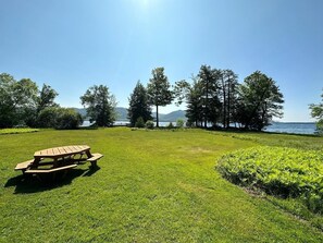 Ein Picknicktisch in einer grünen Wiese mit einem See und Bäumen im Hintergrund.